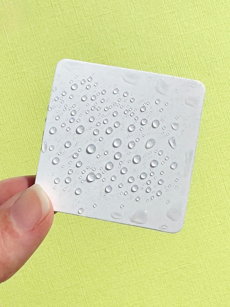 A hand holding a square white foam magnet covered in water droplets against a textured light green background.