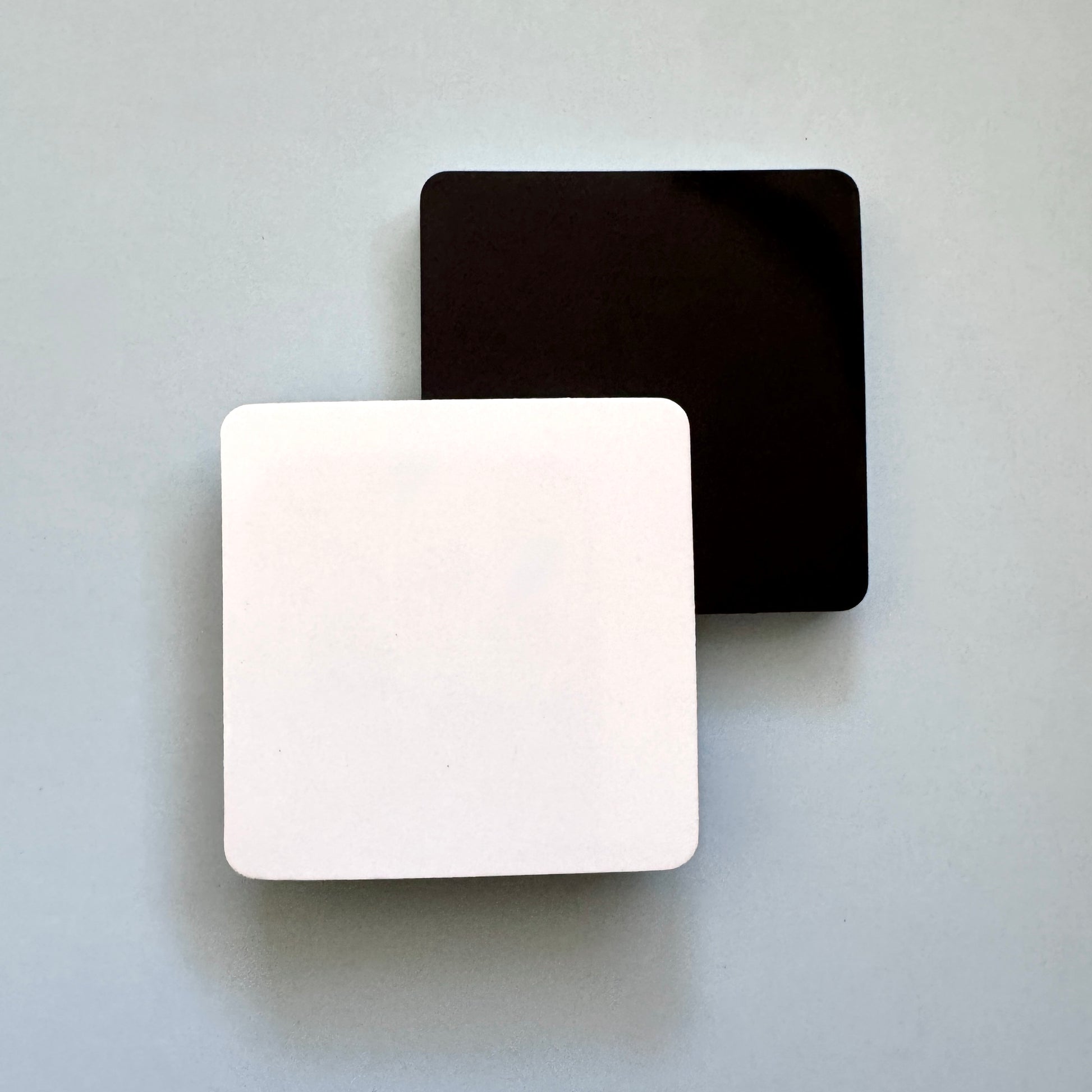 Two square blank magnets on a blue background. One is forward facing with a white front, the other is facing backwards showing the black flat magnet. 