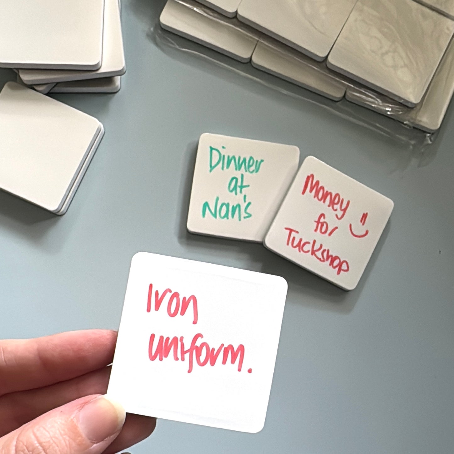Hand holding white magnet with dry-erase marker written on them with the words, iron uniform. Then in the background are more placed with the words, dinner at nan's and money for tuckshop.