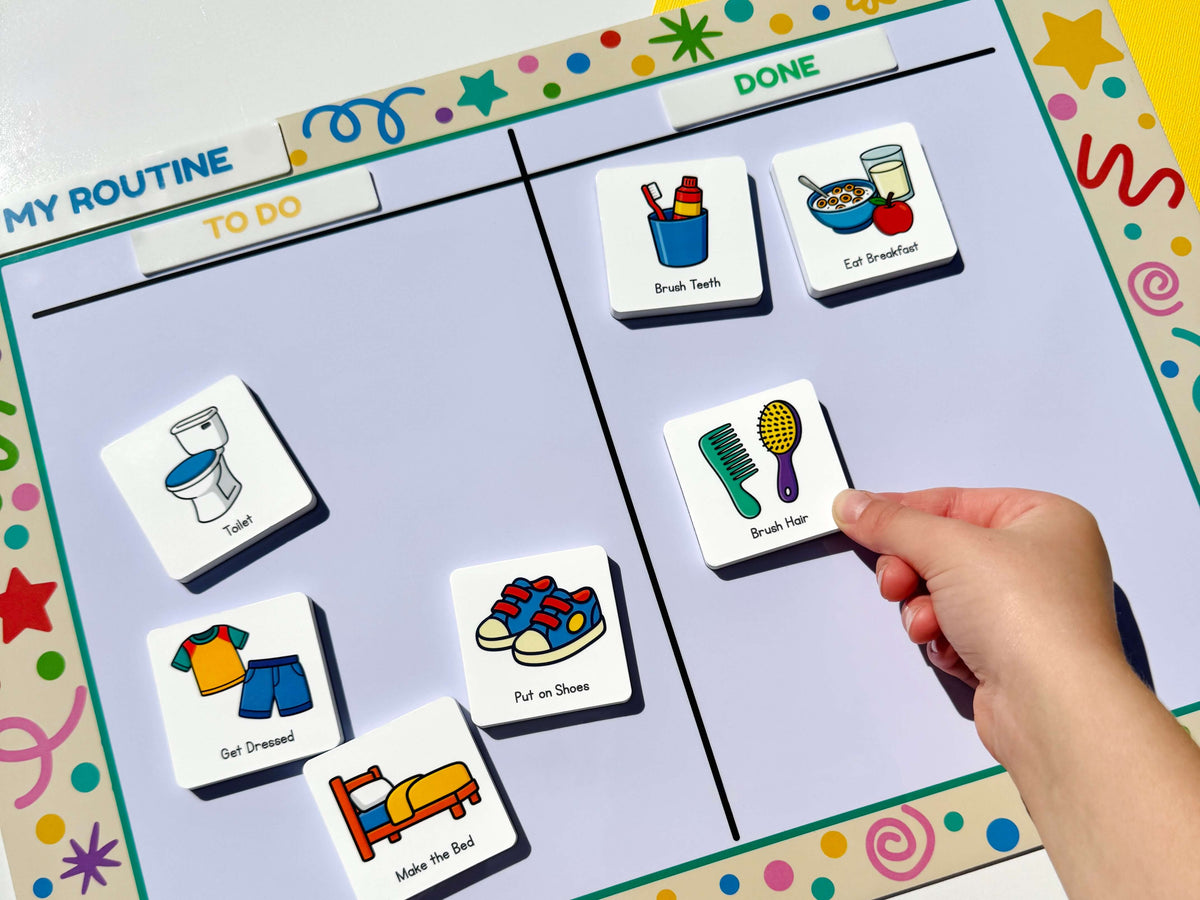 A close-up of a child's hand placing a colourful routine magnet (brush hair) onto a 'My Routine' daily magnetic board. The board features various illustrated magnetics representing daily tasks such as toilet, get dressed, brush hair, and put on shoes, set against a white background.