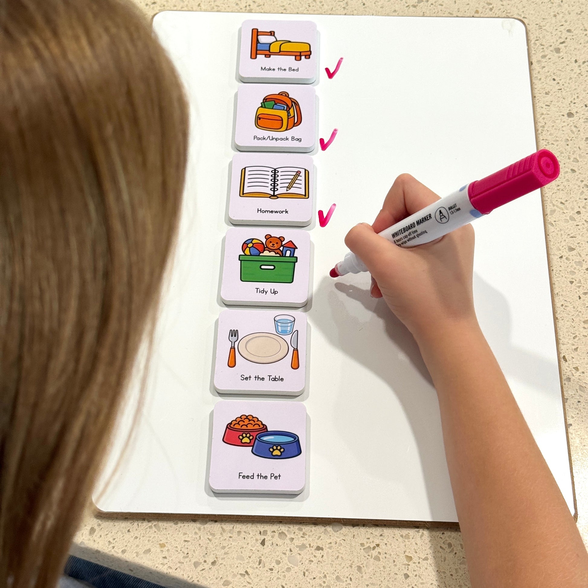 Child using a pink marker to tick beside home task magnets on a whiteboard.