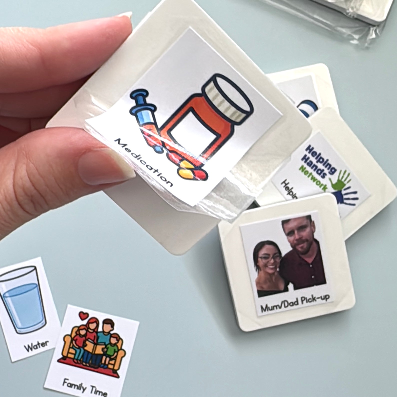 Hand holding a single blank magnet with the sticker medication stuck to its surface being peeled off, with other selection of magnets in the background with various stickers attached. 