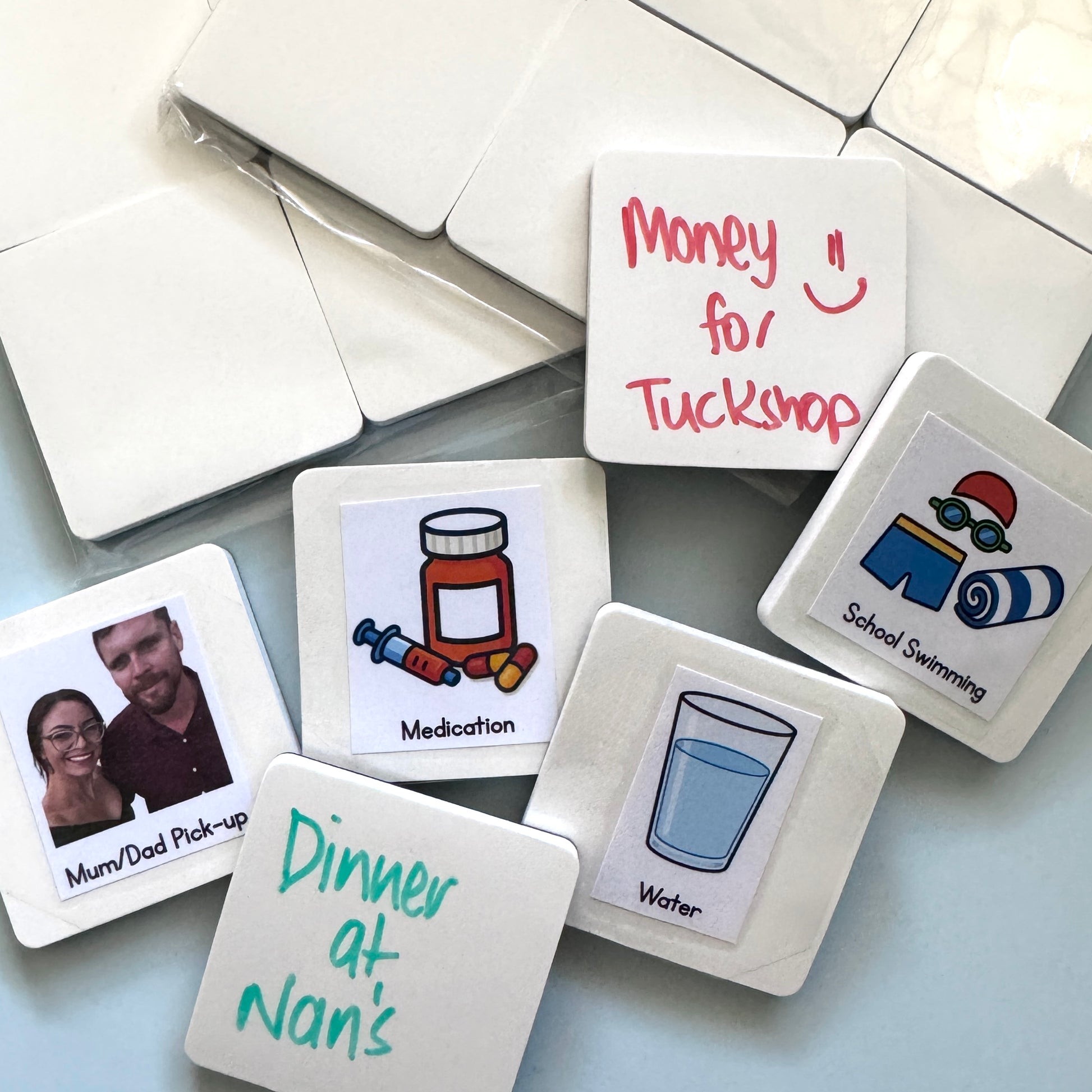A selection of blank white magnets with a selection of either stickers or words written in dry-erase on the surface of them, such as money for tuckshop, school swimming and water. 